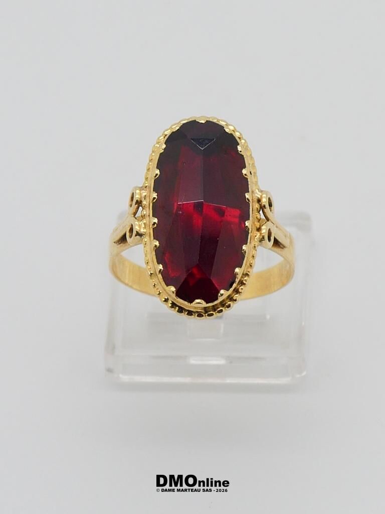 Gold ring with an oval deep red gemstone set in a beaded bezel, on a clear display stand.