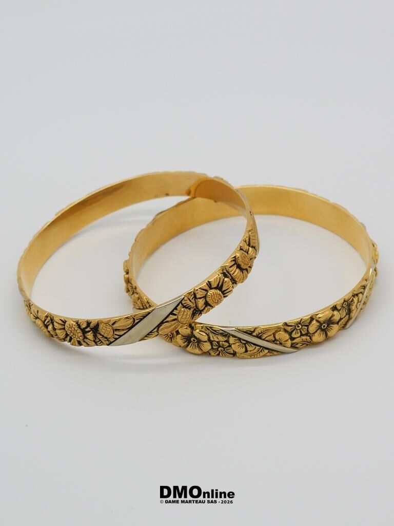 Two gold bracelets with embossed floral patterns, intertwined on a white background, showing a silver diagonal accent.